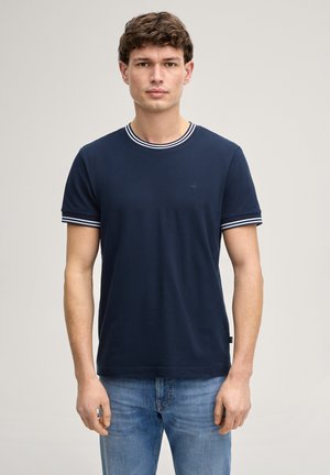 Junger Mann mit kurzen lockigen Haaren, der ein marineblaues T-Shirt mit weißen gestreiften Bündchen und Kragen trägt, kombiniert mit blauen Jeans, steht neutral.