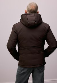 Braune gepolsterte Jacke mit hohem Kragen und verstellbarem Kapuze, mit glatter Textur und Nahtdetails. Getragen über dunkelgrauen Hosen.