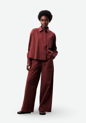 Vrouw die staat met handen in haar zakken, draagt een losvallend bordeauxrood overhemd met knopen en wijd uitlopende broek, samen met zwarte sandalen, tegen een witte achtergrond.