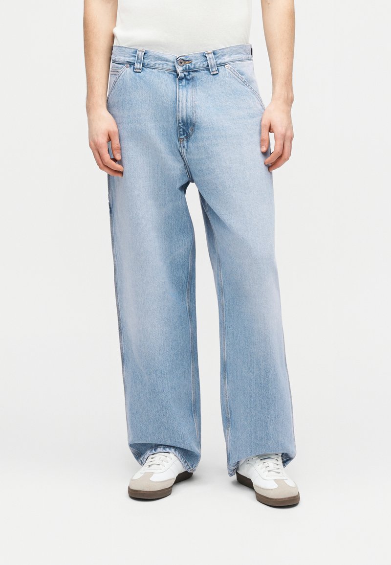 Uomo che indossa jeans azzurri a gamba larga e sneakers bianche, in piedi contro uno sfondo chiaro uniforme.