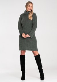 Strickkleid in dunkelgrün, mit hohem Kragen und langen Ärmeln. Kombiniert mit schwarzen knielangen Stiefeln. Weiche Textur mit lockerer Passform.