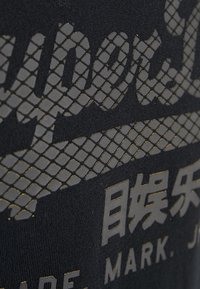 Camiseta negra con un diseño texturizado que presenta patrones de rejilla en relieve en dorado metálico, mostrando el nombre de la marca y caracteres asiáticos.