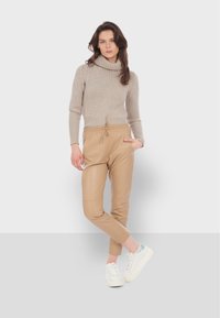 Bežový pletený sveter s vysokým golierom, kombinovaný s pieskovými joggermi z umelého koženého materiálu a bielymi teniskami s svetlomodrým akcentom.