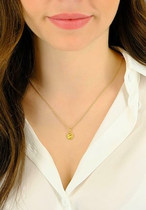 Collana d'oro con un ciondolo rotondo decorato con un motivo di leone, indossata da una persona con una camicia bianca con bottoni.