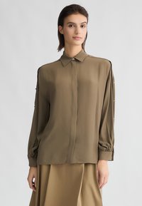 Blusa verde oliva con colletto a bottone, maniche lunghe e polsini arricciati. Presenta un tessuto leggero e morbido, con dettagli accentuati sulle spalle.
