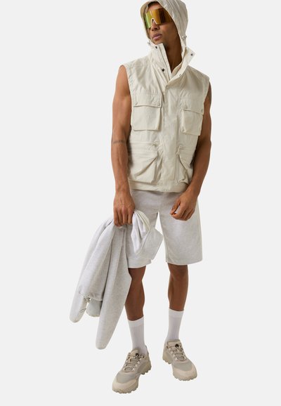 Beige ärmellose Weste mit mehreren Taschen, getragen über einem grauen Hoodie und Shorts. Das Model trägt reflektierende Sonnenbrillen und beige klobige Sneakers.