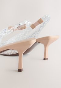 Sandales à talons hauts beiges avec des brides transparentes décorées de perles blanches et de petits strass sur un fond clair.