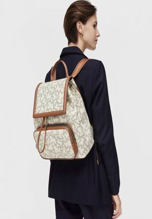 Mochila con una tela texturizada de beige claro que presenta un patrón ondulado, detalles en cuero marrón y correas ajustables. Bolsillo con cremallera en la parte frontal.