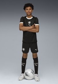Svart fotbollsdräkt med vita detaljer, med en logotyp på bröstet och shorts. Spelaren står med en vit och svart fotboll vid sina fötter.