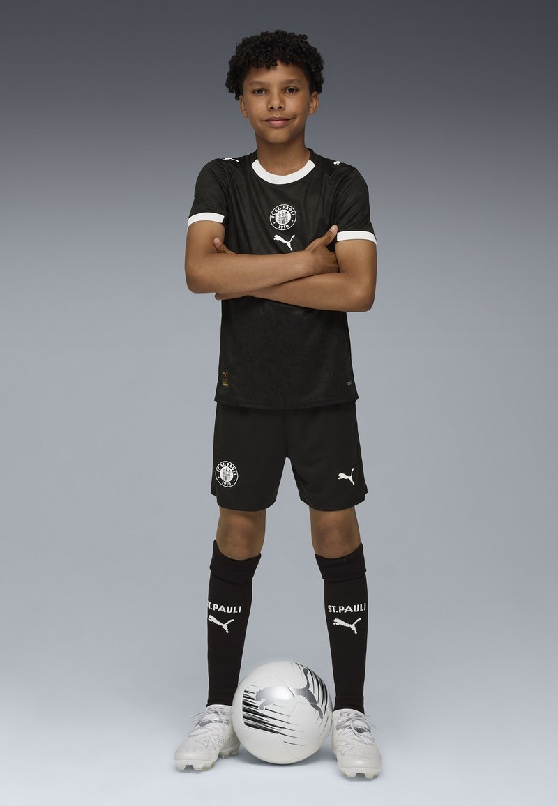 Svart fotbollsdräkt med vita detaljer, med en logotyp på bröstet och shorts. Spelaren står med en vit och svart fotboll vid sina fötter.
