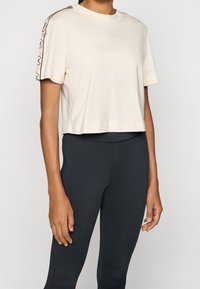 Kobieta ubrana w jasnobeżowy, skrócony T-shirt z taśmą logo na rękawach oraz czarne legginsy o wysokim stanie, stojąca na jednolitym tle.