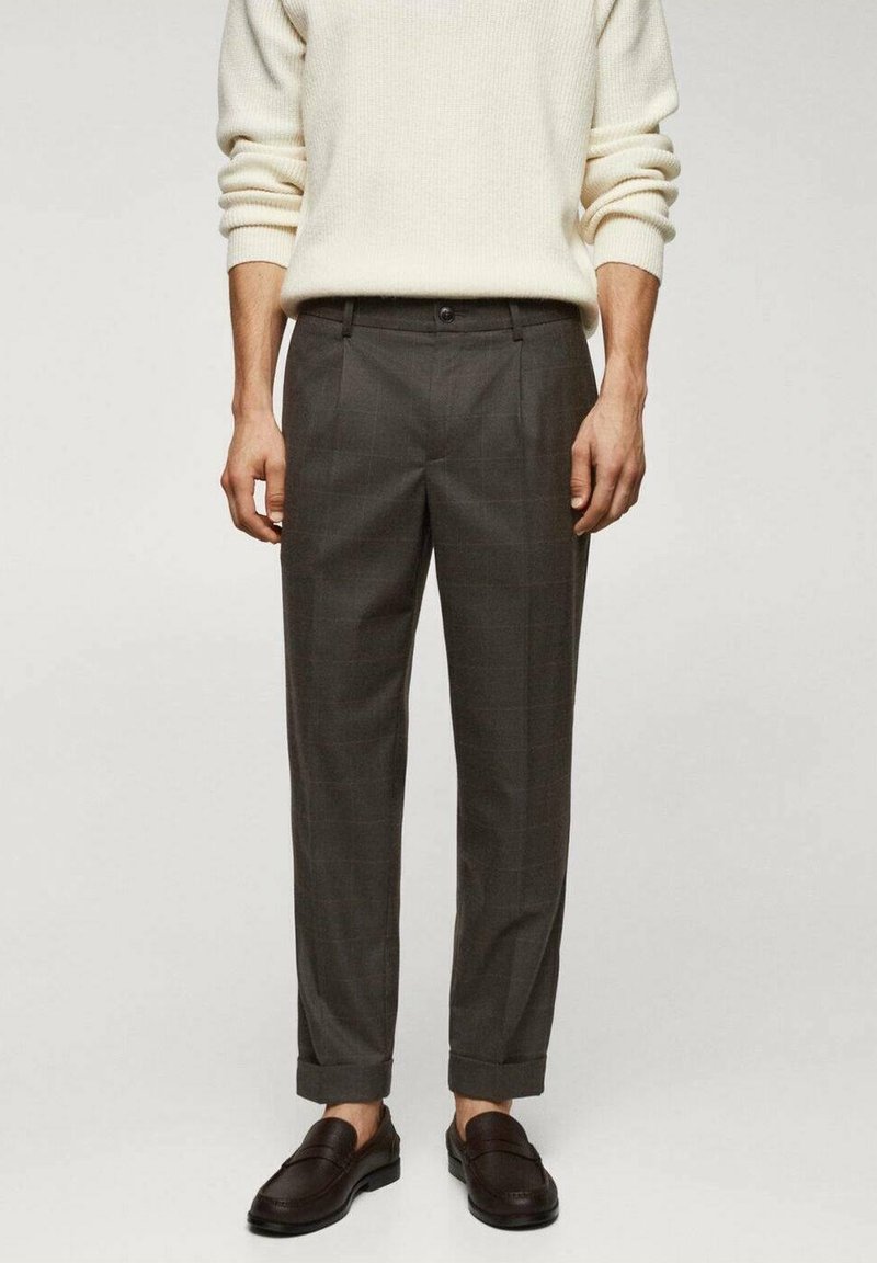 Homme portant un pantalon gris foncé sur mesure avec des revers, associé à un pull en maille crème et des mocassins en cuir marron foncé, debout devant un fond uni.