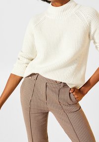 Pull en maille crème avec un col montant et une texture côtelée, associé à un pantalon marron à motif à carreaux avec des lignes verticales et des poches.
