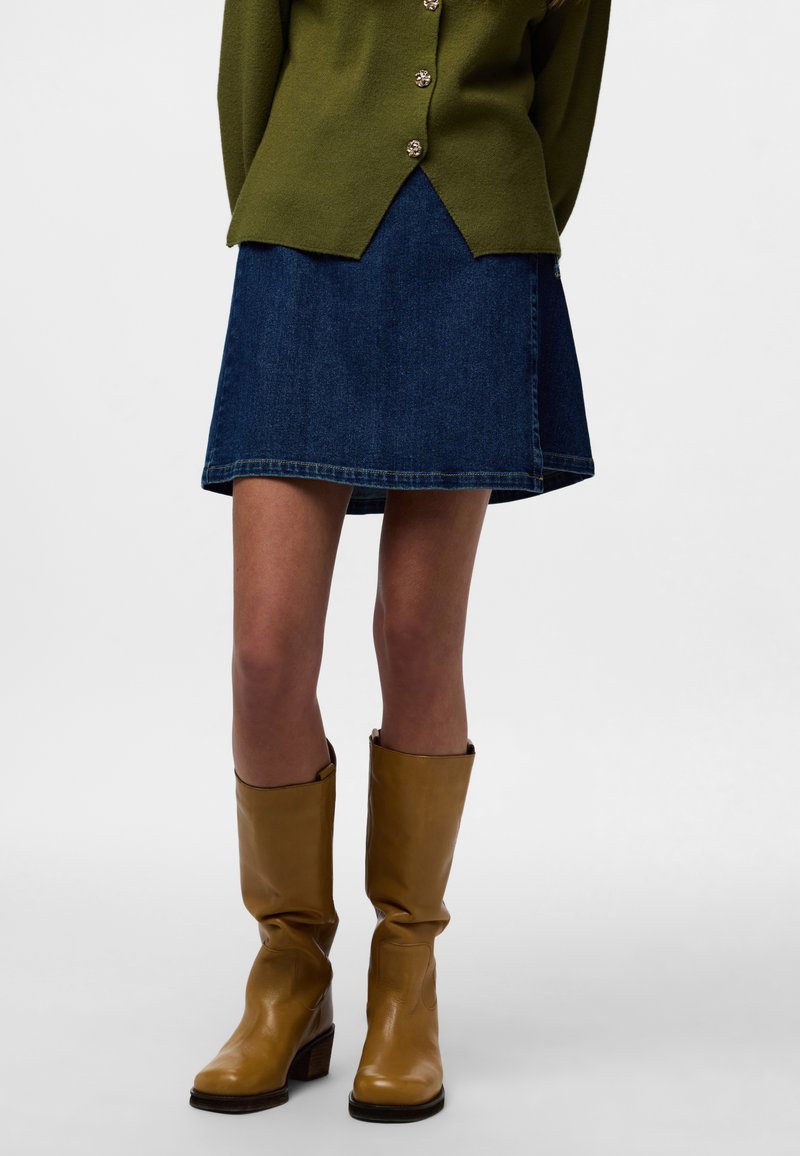 Roheline kardigan nööpidega, sinine teksaseelik ja pruunid nahast põlvikud. Seelikul on laienev serv; saapadel on paks konts.