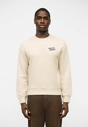 Άνδρας που φοράει κρεμ φούτερ με στρογγυλή λαιμόκοψη και το κείμενο "Maison Kitsuné" στο στήθος, καθώς και σκούρο καφέ παντελόνι, στέκεται μπροστά σε απλό φόντο.