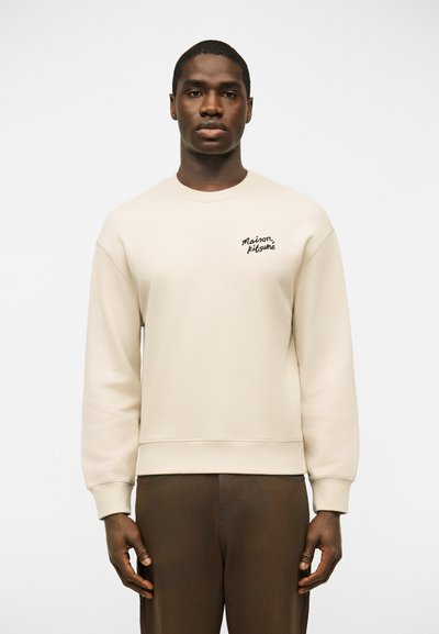 Hombre vistiendo una sudadera crema con cuello redondo y texto "Maison Kitsuné" en el pecho, y pantalones marrón oscuro, posando frente a un fondo liso.