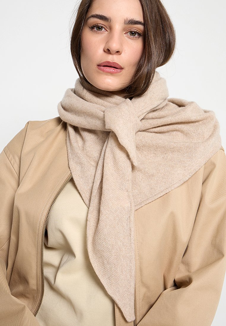 Beige gestrickter Schal um den Hals gewickelt, mit einer dreieckigen Form und weicher Textur, ergänzt von einer hellbraunen Jacke mit Reißverschluss darunter.