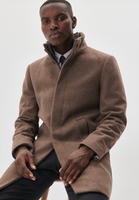 Brauner Wollmantel mit hohem Kragen, ausgestattet mit einer Seitentasche und einem schlanken Design. Der Stoff hat eine weiche Textur und minimale Details.