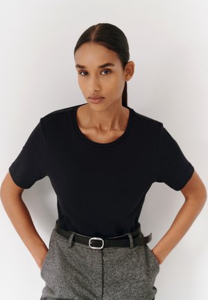 Femme aux cheveux lisses et foncés, vêtue d'un t-shirt noir et d'un pantalon gris avec une ceinture, debout devant un fond blanc les mains dans les poches.