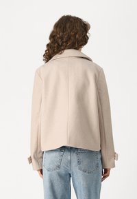 Light beige cropped jacket with a wide collar, long sleeves, and buttoned cuffs. Worn over light blue high-waisted jeans. Soft texture.