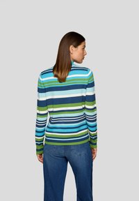 Frau, die von hinten schaut, trägt einen gestreiften Pullover mit blauen, grünen und weißen horizontalen Linien sowie eine blaue Jeans vor einem einfarbigen Hintergrund.