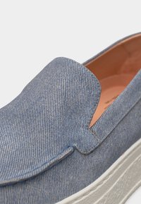 Gros plan sur une chaussure en denim bleu clair à enfiler, avec une semelle en caoutchouc blanche texturée et une doublure intérieure en cuir tanné.