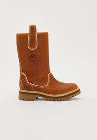 Bruna nahkasaapas keskikorkealla varrella, vetolongalla, vaaleilla ompeluyksityiskohdilla, tekstuuripohjalla ja kohokuvioidulla pyöreällä puulogolla sivussa.