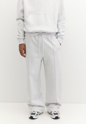 Personne portant un pantalon de survêtement gris clair avec taille à cordon et des baskets blanches et noires, debout devant un fond blanc uni.