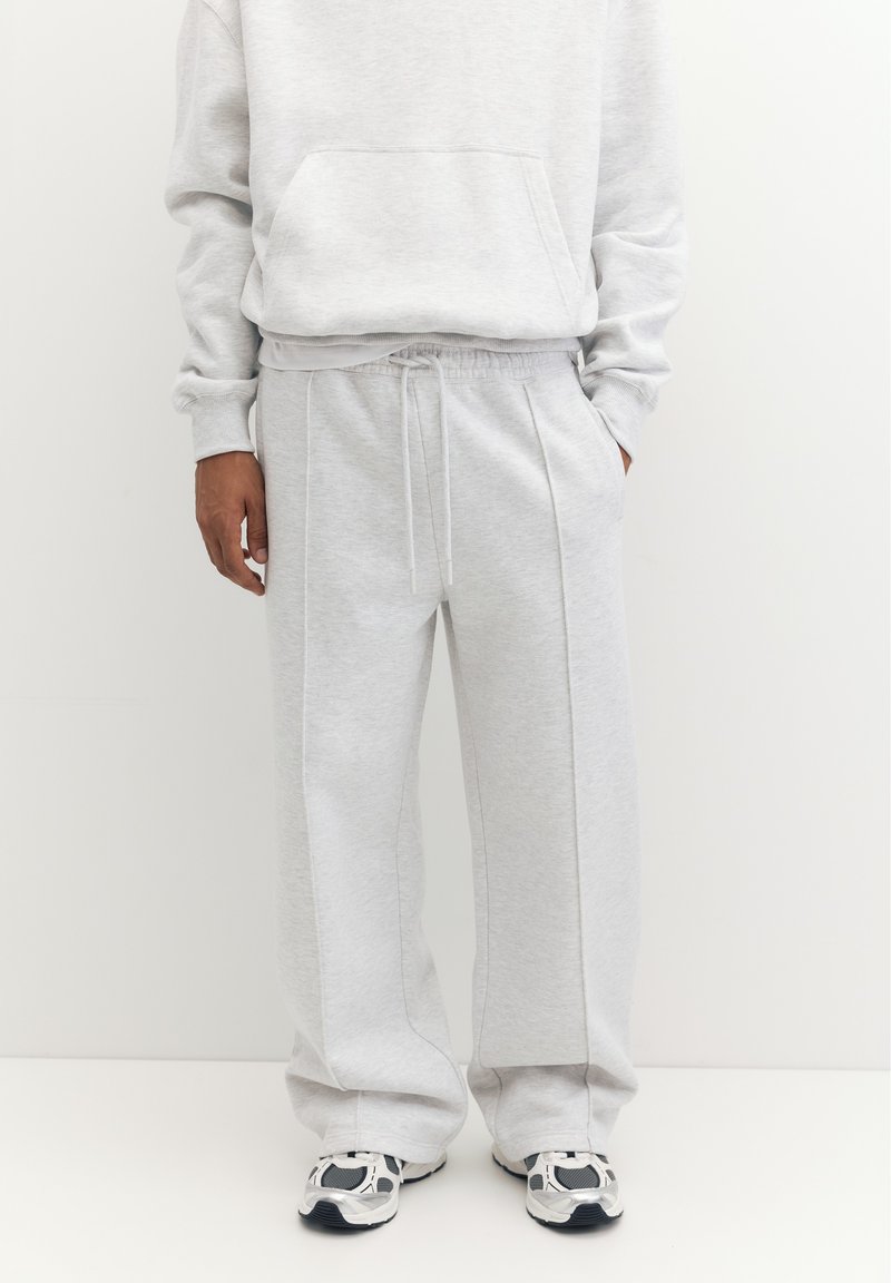 Personne portant un pantalon de survêtement gris clair avec taille à cordon et des baskets blanches et noires, debout devant un fond blanc uni.