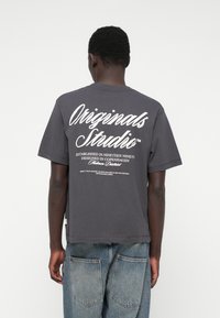 T-shirt de algodão cinza escuro com mangas curtas, apresenta grande texto branco nas costas, decote redondo e barra reta. Ajuste relaxado.