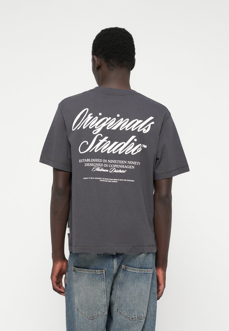 T-shirt de algodão cinza escuro com mangas curtas, apresenta grande texto branco nas costas, decote redondo e barra reta. Ajuste relaxado.