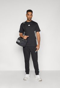 Tricou sport negru cu dungi albe, pantaloni de trening negri cu accente verzi și o geantă de sală neagră cu un logo alb. Pantofi albi.