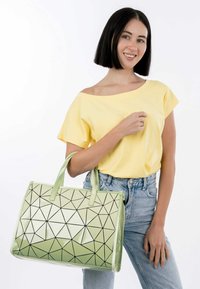 Borsa tote geometrica verde con finitura lucida, motivi triangolari neri e manici abbinati. Modella che indossa una maglietta gialla e jeans.