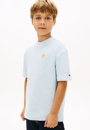 Blonder Junge, der nach vorne blickt und ein hellblaues, strukturiertes Kurzarmhemd mit kleinen aufgestickten Logos auf Brust und Ärmel trägt.