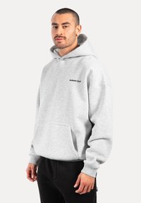 Hoodie grigio in cotone con tasca frontale, polsini a coste e logo ricamato sul petto. Il modello indossa pantaloni della tuta neri.