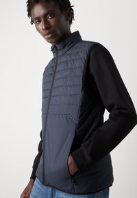 Colete acolchoado azul-marinho com gola alta, textura acolchoada e mangas longas pretas. Possui fecho de correr e bolsos laterais para maior praticidade.