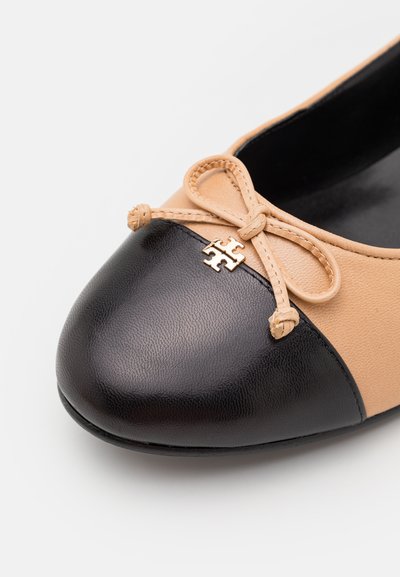 Ballerine tan et noire avec un bout arrondi, dessus en cuir, nœud décoratif et un petit logo doré sur le bout.