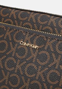 Zbliżenie czarnego portfela o teksturowanej powierzchni z brązowym wzorem logo CK i złotym napisem Calvin Klein obok złotego zamka błyskawicznego.