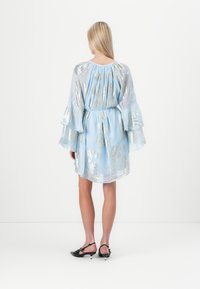 Vestido curto azul claro com padrões de folhas metálicas prateadas, mangas abertas e detalhe franzido nas costas, combinado com sapatos slingback pretos.