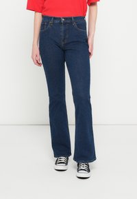 Vaqueros acampanados azules hechos de mezclilla, con bolsillos delanteros, cierre de botón y cremallera, y un corte clásico. Combinados con zapatillas negras.