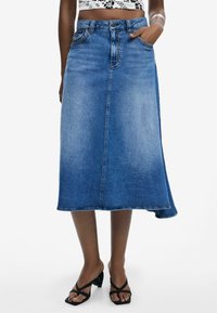 Jeans-A-Linienrock in Mittelblau mit hoher Taille, Fronttaschen und rohem Saum. Begleitet von schwarzen Riemchensandalen.