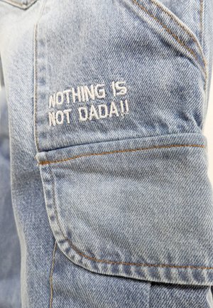 Blaue Denim-Stoff mit Stickdetails. Mit gesticktem Text: "NICHTS IST NICHT DADA!!", der auffällig im Taschenbereich platziert ist.