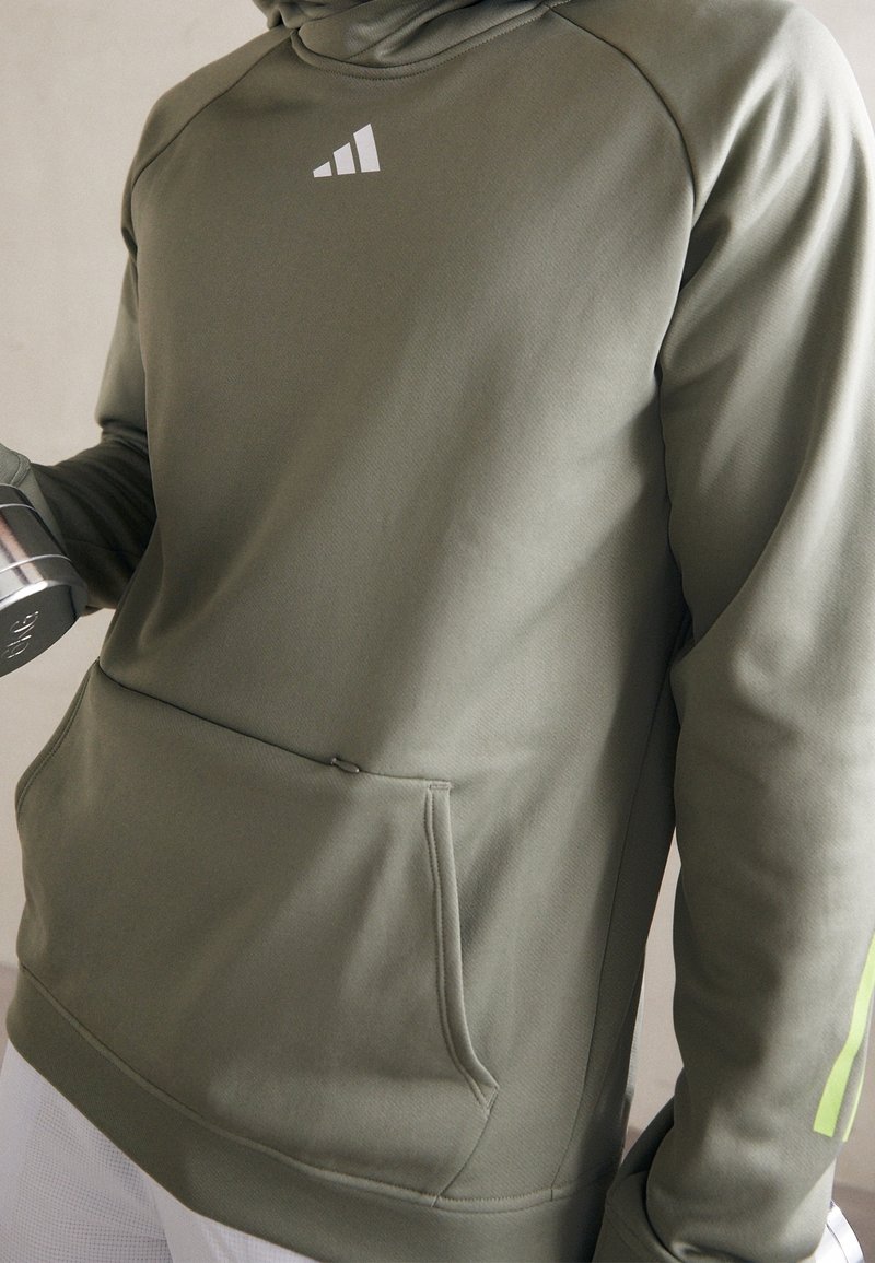 Personne portant un sweat à capuche Adidas vert olive tenant un haltère argenté dans une main, montrant le haut du torse et une partie des manches.