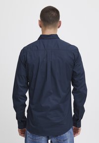 Maglietta a maniche lunghe blu navy in cotone, con colletto classico, dettaglio di piega sul retro e polsini con bottone. Vista da dietro.