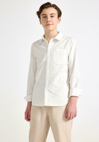 Biała koszula z długim rękawem wykonana z lekkiego materiału, z zapięciem na guziki, wyostrzonym kołnierzykiem i kieszenią na piersi. Noszona z beżowymi spodniami.