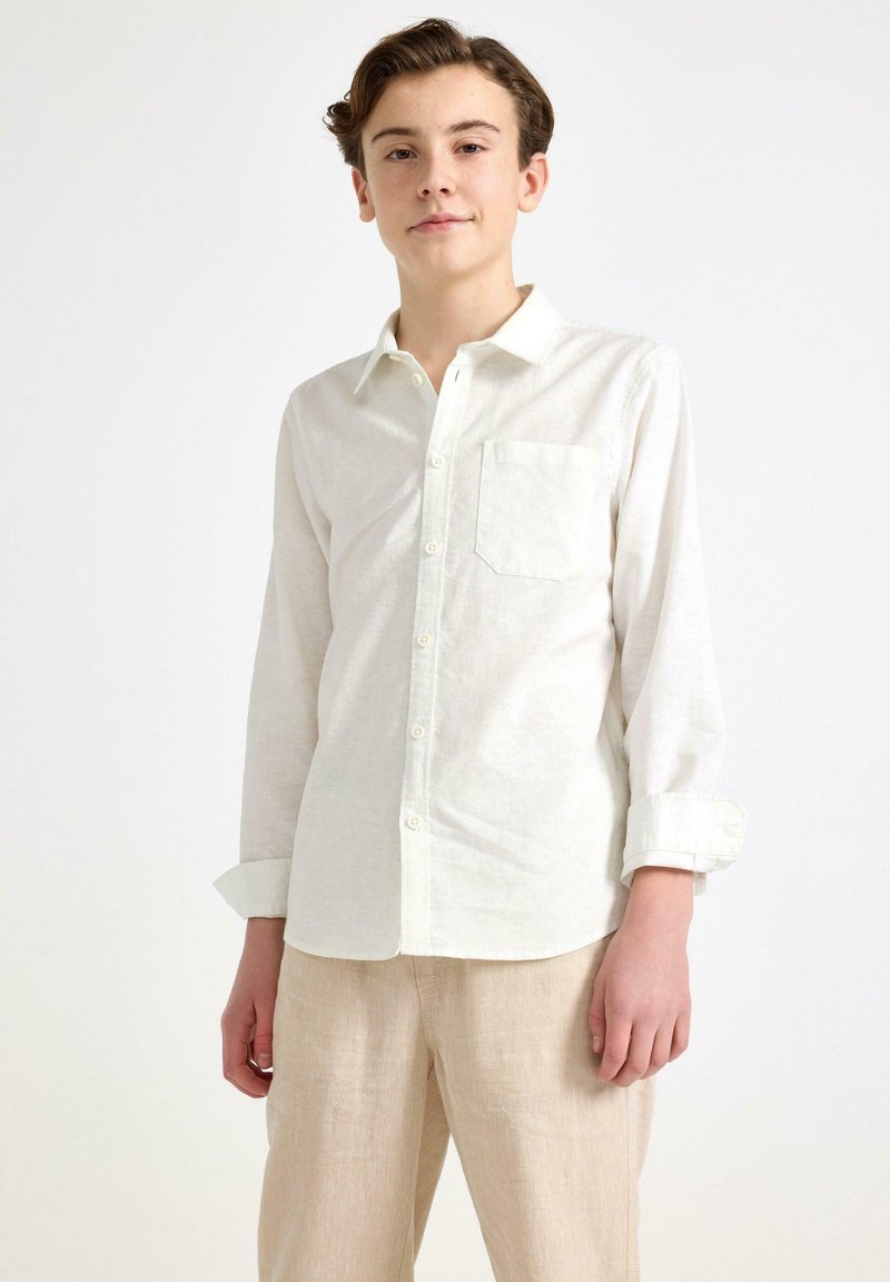 Biała koszula z długim rękawem wykonana z lekkiego materiału, z zapięciem na guziki, wyostrzonym kołnierzykiem i kieszenią na piersi. Noszona z beżowymi spodniami.