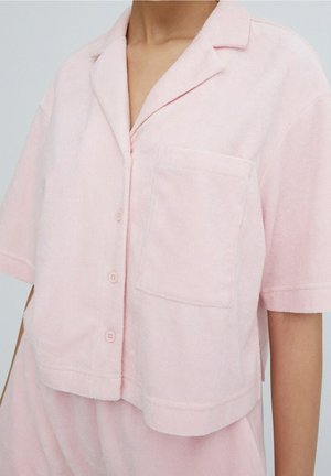 Haut de pyjama à manches courtes en toile éponge de couleur rose clair. Il présente un col à revers, une poche avant et cinq boutons de fermeture.
