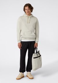 Felpa grigia chiara con coulisse, tasca a marsupio, indossata con pantaloni sportivi neri e sneakers beige. Una borsa tote resistente è tenuta in una mano.