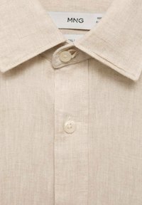 Camisa de lino beige con cuello clásico, botón frontal con dos botones y una tela texturizada. Marca MNG visible en el interior del cuello.