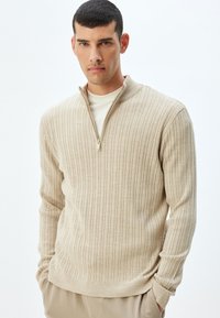 Suéter de canalé beige con cuello con cremallera, que presenta una tela texturizada y un diseño ajustado. Destacado por las mangas largas y un corte slim.